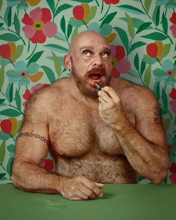 A shirtless, bald adult male with a beard applies red lipstick in front of a colorful floral wallpaper and green table, looking up.